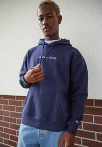 Sudadera con capucha azul marino, con un bolsillo frontal y el logotipo bordado de "Tommy Jeans". Presenta cordones ajustables y puños de canalé. Se lleva con jeans de color azul claro.