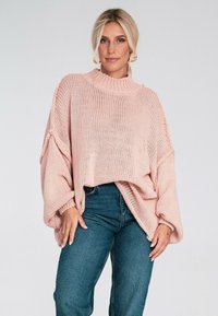 Jasnoróżowy, dziany sweter z wysokim dekoltem, w oversize'owym kroju i szerokimi rękawami; zestawiony z niebieskimi dżinsami.