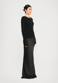 Mulher de lado vestindo um cardigan preto com botões em tecido canelado e uma saia longa preta de seda contra um fundo branco liso.