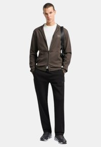 Felpa con zip marrone sopra una maglietta bianca, abbinata a pantaloni neri. Il modello indossa sneakers nere e porta una borsa nera. Tessuto morbido, vestibilità rilassata.