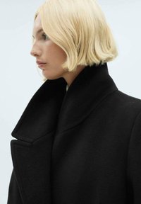 Cappotto di lana nera con colletto alto e ampi risvolti, che mostra una texture morbida e linee pulite. Profilo laterale del modello visibile.