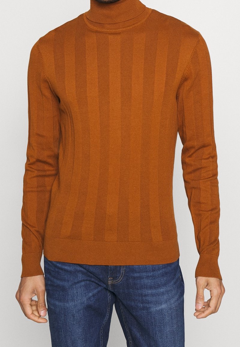 Homme portant un pull à col roulé côtelé ajusté de couleur rouille orangée et un jean bleu, debout devant un fond clair uni.