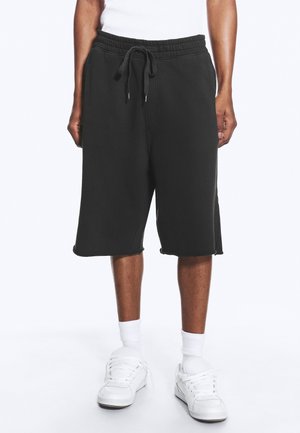 Person iført sorte knælange shorts med snor, hvide sokker, hvide sneakers og en hvid top mod en ensfarvet baggrund.