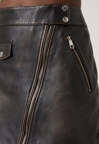 Svart läderkjol med en sliten finish, asymmetrisk frontdragkedja, tryckknappar och en sidoficka med silverfärgad dragkedja.