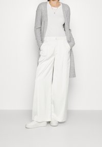 Hellgraue Strickjacke, weißes eng anliegendes Oberteil, weiße, weit geschnittene Hose mit vorderen Falten und weiße Sneakers. Minimalistisches Design, glatte Texturen.