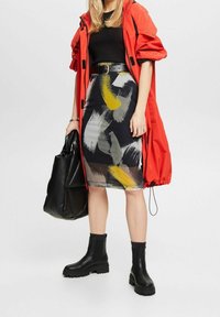 Femme portant une veste rouge, un haut noir côtelé, une jupe crayon à motif abstrait, une ceinture noire et des bottes, tenant un grand sac à main noir.