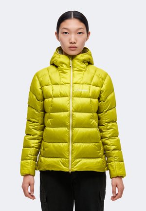 Une femme porte une doudoune à capuche vert citron vif, fermée par une fermeture éclair sur le devant, ainsi qu’un pantalon noir, sur un fond clair uni.