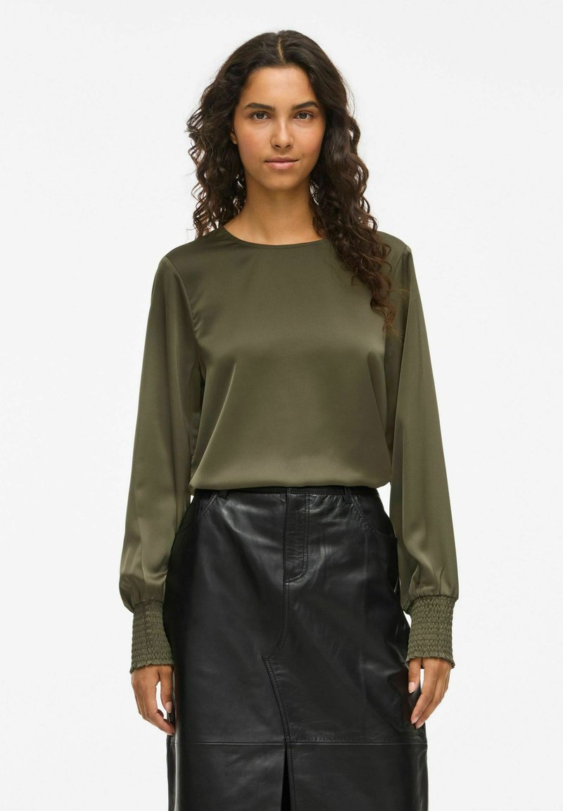 Vrouw met lang krullend haar, gekleed in een olijfgroene satijnen blouse en een hooggepaste zwarte leren rok tegen een witte achtergrond.