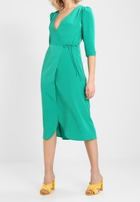 Green wrap dress with three-quarter sleeves, cinched waist tie, and front slit. Paired with yellow block-heeled sandals. Smooth texture.