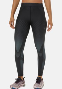 Leggings noirs taille haute avec des motifs géométriques gris, offrant une texture lisse et un ajustement serré. Associés à des chaussures de course violettes claires.
