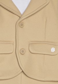 Beige kavaj med texturerad väv, två framfickor, två knappar och en vit logotyp på vänstra fickan.