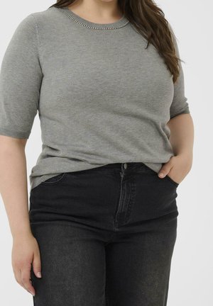 Frau trägt ein graues kurzärmeliges Stricktop mit perlenverziertem Halsausschnitt und hoch taillierte schwarze Jeans, Hand in der Tasche.