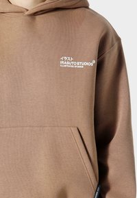 Brun hættebluse lavet af blød stof med en frontlomme. Har et hvidt tekstlogo på venstre side og et mærke på kanten.