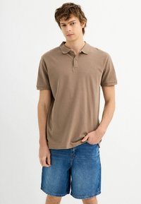 Camisa polo de algodón marrón con cuello, mangas cortas y un pequeño logo, combinada con pantalones cortos de mezclilla. La tela parece suave y ligera.