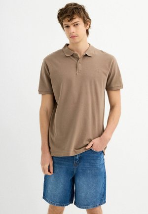 Camisa polo de algodón marrón con cuello, mangas cortas y un pequeño logo, combinada con pantalones cortos de mezclilla. La tela parece suave y ligera.