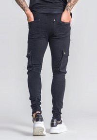 Pantalones cargo de mezclilla negra con corte ajustado, bolsillos laterales tipo cargo y bolsillos traseros de parche. Estilizados con una cinturilla ajustada y un diseño de largo hasta el tobillo.