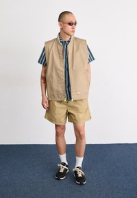 Beige ermeløs vest over en blå- og hvitstripet skjorte, paret med beige shorts og svarte joggesko; har sidelommer og grønne detaljer.