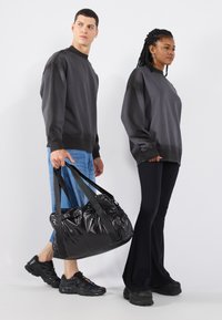 Dva modela nose tamnosive oversized dukserice. Jedan drži sjajnu crnu torbu sa crnim kaiševima. Drugi nosi crne široke pantalone i patike.