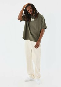 Camiseta oversized de color verde oliva con un gráfico en el pecho, combinada con pantalones de algodón color crema y zapatillas blancas.