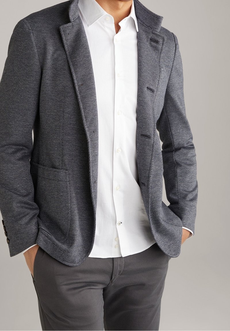 Blazer grigio testurizzato con rever a notches e due tasche frontali, indossato sopra una camicia bianca e pantaloni grigi. Sottile motivo a spina di pesce.