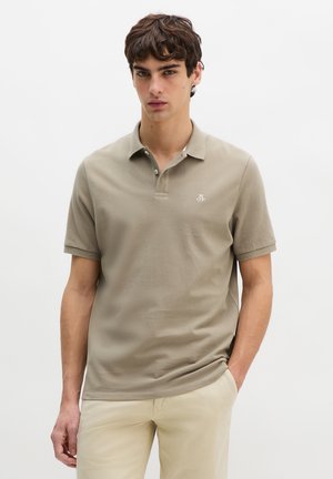 Νέος άνδρας που φοράει ένα ταουπέ κοντομάνικο πόλο με μικρό λευκό λογότυπο και μπεζ παντελόνι, στέκεται με το ένα χέρι στην τσέπη.