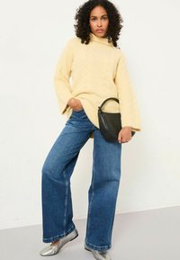 Cremfarbener Kabelstrickpullover mit Glockenärmeln, kombiniert mit weiten blauen Jeans. Metallisch silberne Ballerinas und eine kleine schwarze Handtasche runden den Look ab.