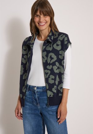 Uśmiechnięta kobieta ubrana w biały shirt z rękawem do łokcia, niebieskie dżinsy i granatową kamizelkę z zielonym wzorem w cętki.