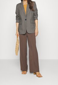 Blazer gris à carreaux avec deux poches devant, associé à un haut marron et un pantalon ample assorti. Sac tissé beige à la main, sandales à brides marron.