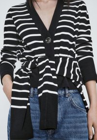 Czarny i biały kardigan w paski z głębokim dekoltem w serek, zapinany na guziki, z wiązanym paskiem, w połączeniu z niebieskimi dżinsami.