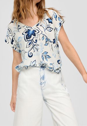 Frau trägt eine weiße Bluse mit blauen Blumenmustern und hoch taillierte weiße Jeans mit blauer Naht, posiert mit einem ausgestreckten Arm.