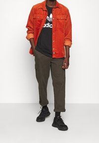 Oranžová kordurová košile, černé tričko s bílým logem, olivové cargo kalhoty a černé tenisky. Má ohrnuté rukávy a boční kapsy.