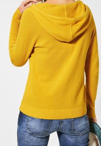Gelbe strukturierte Kapuzenjacke mit Kapuze, geripptem Saum und langen Ärmeln, kombiniert mit blauen Jeans, die über Taschen auf der Rückseite und dezente Nähte verfügen.