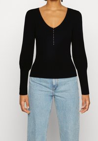 Pull noir à côtes avec un décolleté en V profond et des manches longues, comportant une rangée de boutons métalliques sur le devant et un design ajusté.