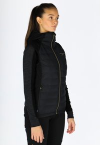 Gilet imbottito nero con chiusura zip e cappuccio, abbinato a maniche lunghe nere. Presenta dettagli in oro e tasche laterali con una texture liscia.