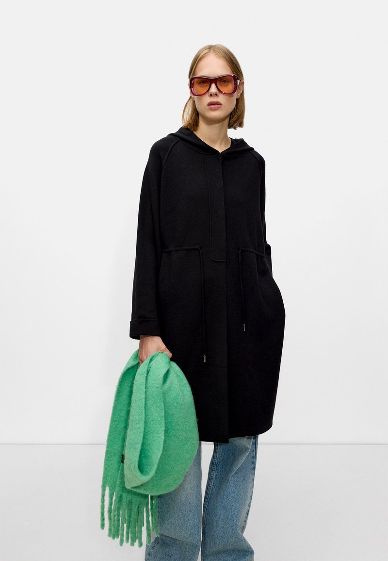 Manteau en polaire noir avec une capuche, des poches avant et une coupe décontractée. Tenant une écharpe verte texturée avec des franges. Lunettes de soleil roses.
