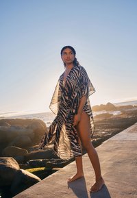 Durchsichtiger, langer Kimono mit einem schwarz-braunen Zebramuster, ausgestattet mit einem tiefen V-Ausschnitt und weiten Ärmeln, die sich an der Figur am Strand sanft entlangfließen.