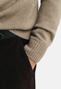 Béžový žebrovaný pletený svetr s elastickými manžetami, spárovaný s tmavohnědými manšestrovými kalhotami. Na pase je viditelný černý pásek.