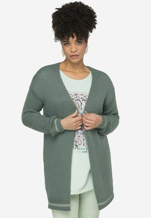 Grøn strikket cardigan med V-hals, lange ærmer og ribbede manchetter. Båret over en lyseblå grafisk t-shirt med blomsterdetaljer.