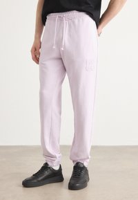 Lyse lilla joggerbukser med snor i taljen, elastiske manchetter og broderet "HUGO" logo. Båret med sorte sneakers på et neutralt gulv.