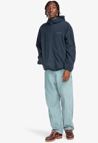 Mørkeblå fleece zip-up jakke med hætte, der har en brystlomme. Parret med lyseblå afslappede jeans og brune snøresko.