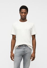 Νεαρός άνδρας που φοράει λευκό κοντομάνικο T-shirt με σχέδια και ανοιχτό γκρι τζιν, στέκεται μπροστά σε απλό λευκό φόντο.