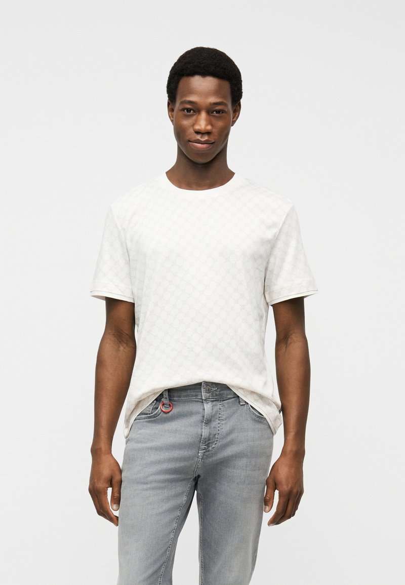 Νεαρός άνδρας που φοράει λευκό κοντομάνικο T-shirt με σχέδια και ανοιχτό γκρι τζιν, στέκεται μπροστά σε απλό λευκό φόντο.