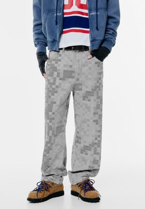 Grau pixelierte Musterhose aus locker sitzendem Stoff. Kombiniert mit braunen strukturierten Schuhen und blauen Schnürsenkeln, ausgestattet mit Fronttaschen.