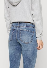 Jeans de denim azul com um design ajustado, apresentam cinco bolsos, costura em contraste e um remendo de couro castanho na parte de trás da cintura.