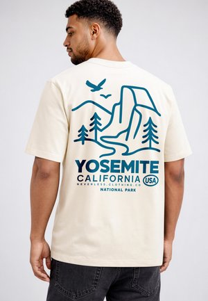 Man wearing beige t-shirt with blue Yosemite National Park design, including mountains, trees, birds, and text on the back.