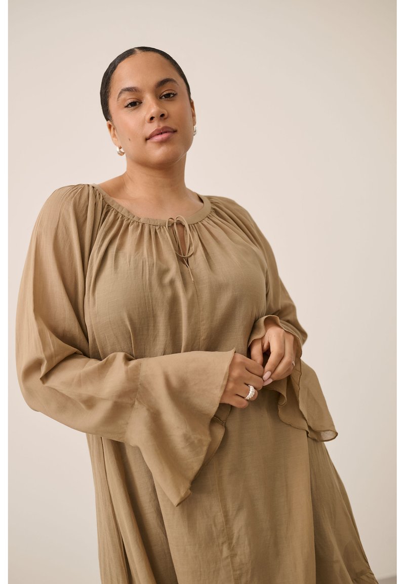 Femme aux cheveux foncés attachés, portant une robe beige ample à manches longues avec des poignets évasés, les mains près de la taille contre un fond uni.