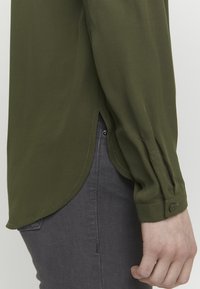 Olive green long-sleeve blouse with a rounded hem, buttoned cuff, and smooth fabric; paired with dark gray pants.