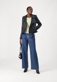 Crna kožna biker jakna s patentnim zatvaračima, zeleni pletivasti top i široke plave traperice. Dodatci uključuju malu crnu torbicu i štikle.