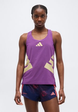 Femme portant un débardeur violet Adidas et un short Adidas bleu marine avec des motifs rouges, debout devant un fond blanc uni.