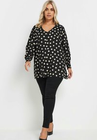 Svart blus med prickar, V-hals och långa puffärmar. Tillverkad av lättviktig tyg och har en avslappnad passform för komfort. Kombineras med svarta byxor.